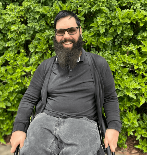 Photo of Nick Schumi seated in his wheelchair with a hedge in the background