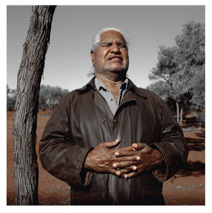 A photo of Yami Lester, outside, with his hands clasped in front of him, his eyes closed, and his head tilted back with his face towards the sky